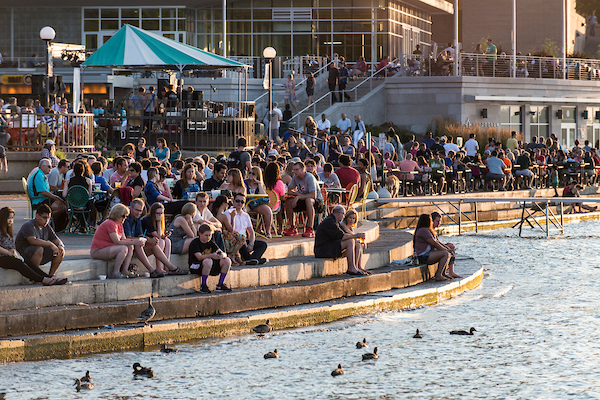 People sitting along the curving cement steps that hug the Lake Mendota shoreline and Memorial Union Terrace as the sun sets on a summer evening.